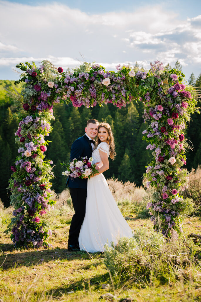 Bride and groom hug and smile to look at the camera in front of the flowered arch with purple flowers.