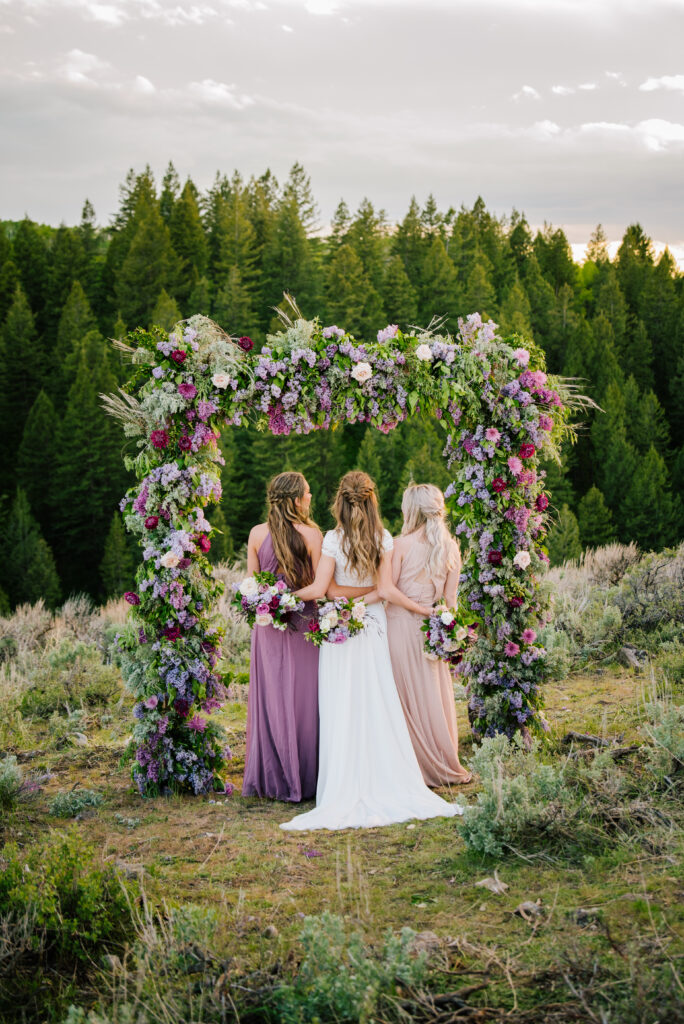 Bride and Bridesmaid's turn their back to the camera to face an elaborate purple flower arch after grand Teton wedding!