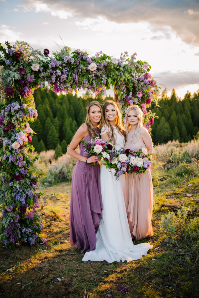 Bride with purple flowered arch holds her purple flowers next to her bridesmaids in purple tones after their mountain colorful grand teton national park wedding.
