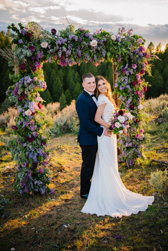 Bride and groom stand in front of purple flower arch and smile at the camera after their Grand Teton wedding.