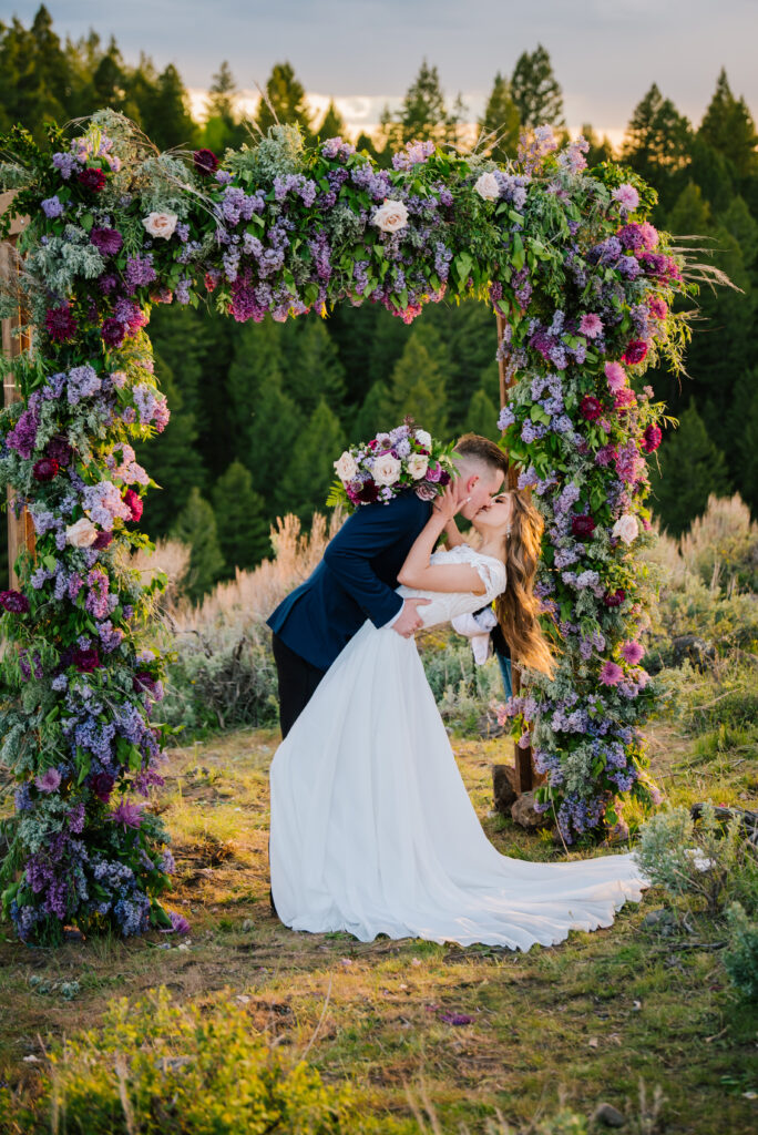 Jackson Hole wedding groom dips bride to kiss her at the end of their mountain ceremony with thick pine trees and a beautiful purple flower arch.