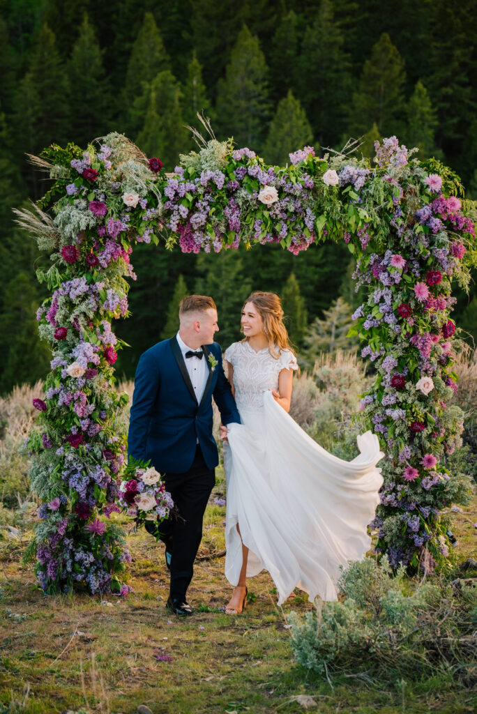 Grand Teton national park wedding bride and groom walk away from purple flowered arch. Groom is carrying the brides bouquet and bride is laughing holding her flowing dress.