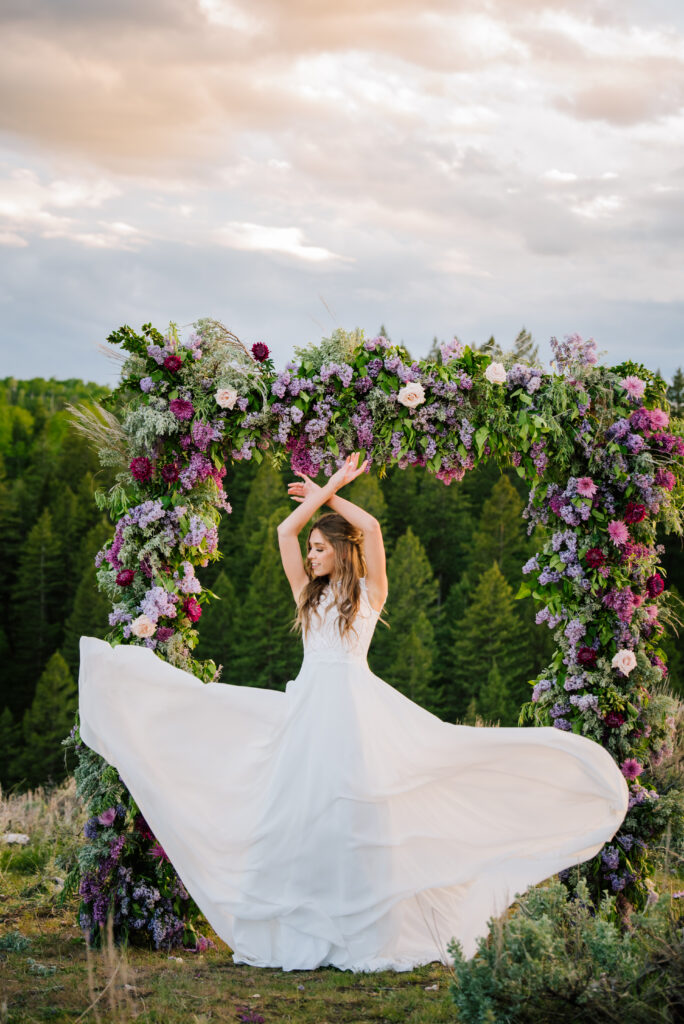 Bride standing in front of a purple and lilac flowered arch. She raises her arms above her head as her dress flows in the wind.