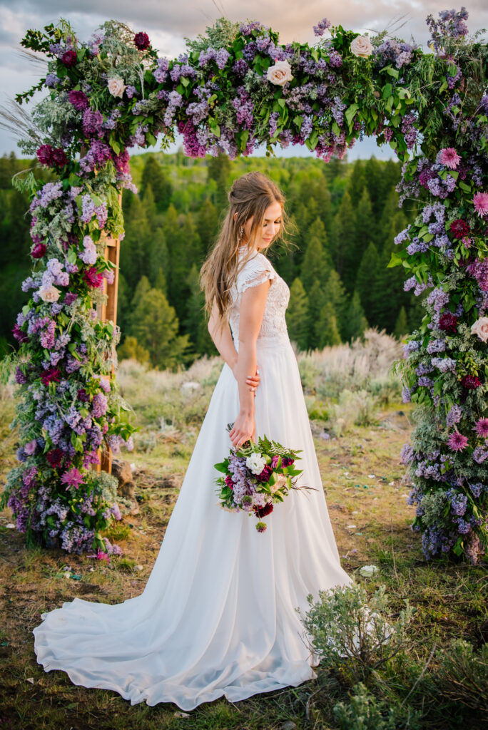 Bride looks down her arm towards her bouquet. Behind her is a arch that is covered in purple toned flowers in Grand Teton National Park.