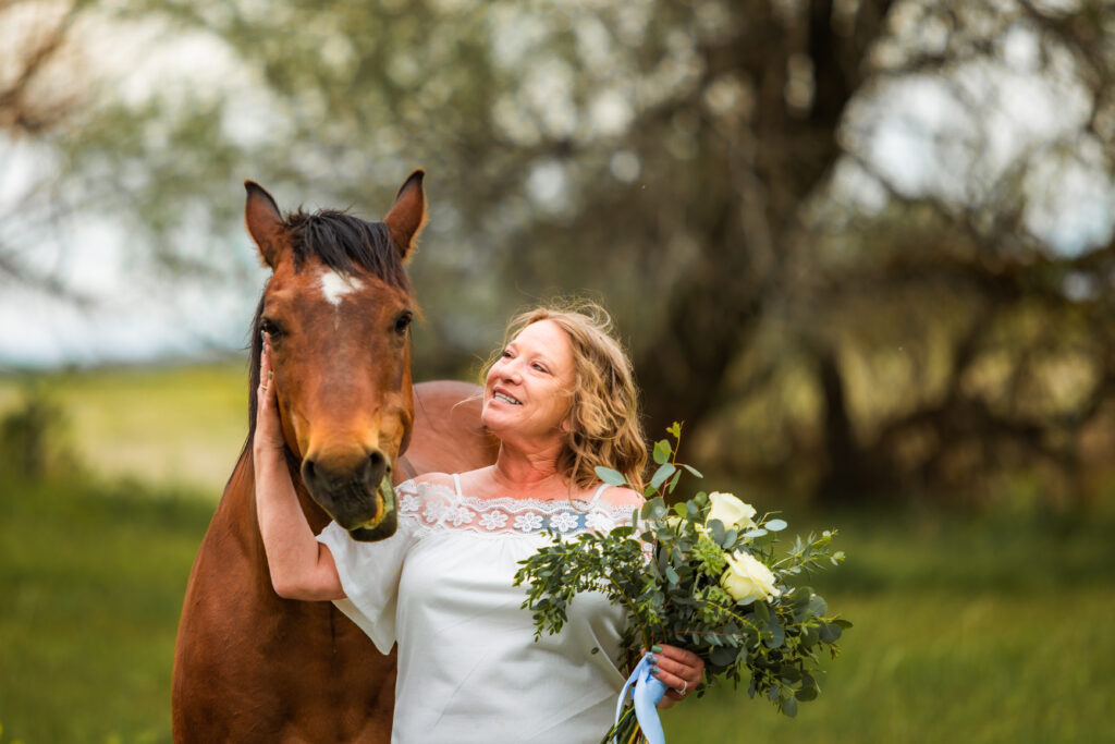 Bride in white shirt holds horse and smiles with one hand while holding her bouquet in the other hand.