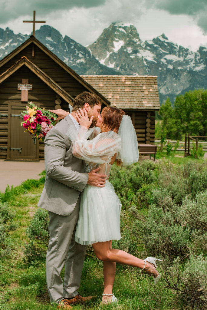 chapel of transfiguration elopement couple embrace in kiss after their jackson hole elopement. Bride is wearing a short dress and veil. 