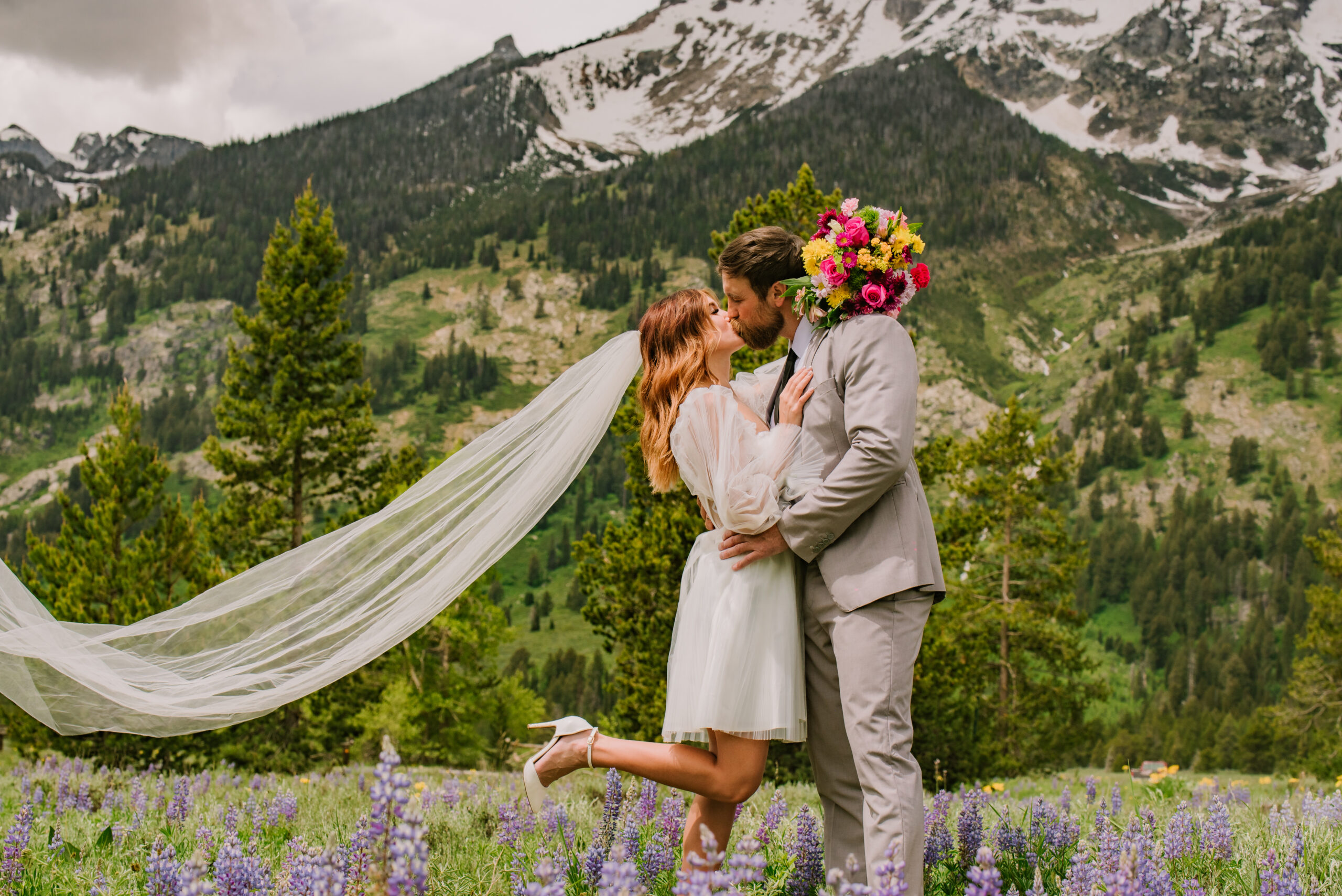 Bride in short dress and long veil kisses her groom in front of green mountain. Her bright colored bouquet is behind his head and her foot is lifted up.