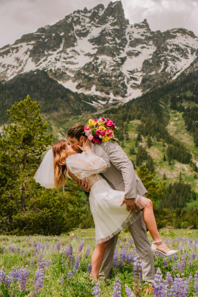 groom dips and kisses bride while holding her leg in grand teton national park in a field of wildflowers. 
