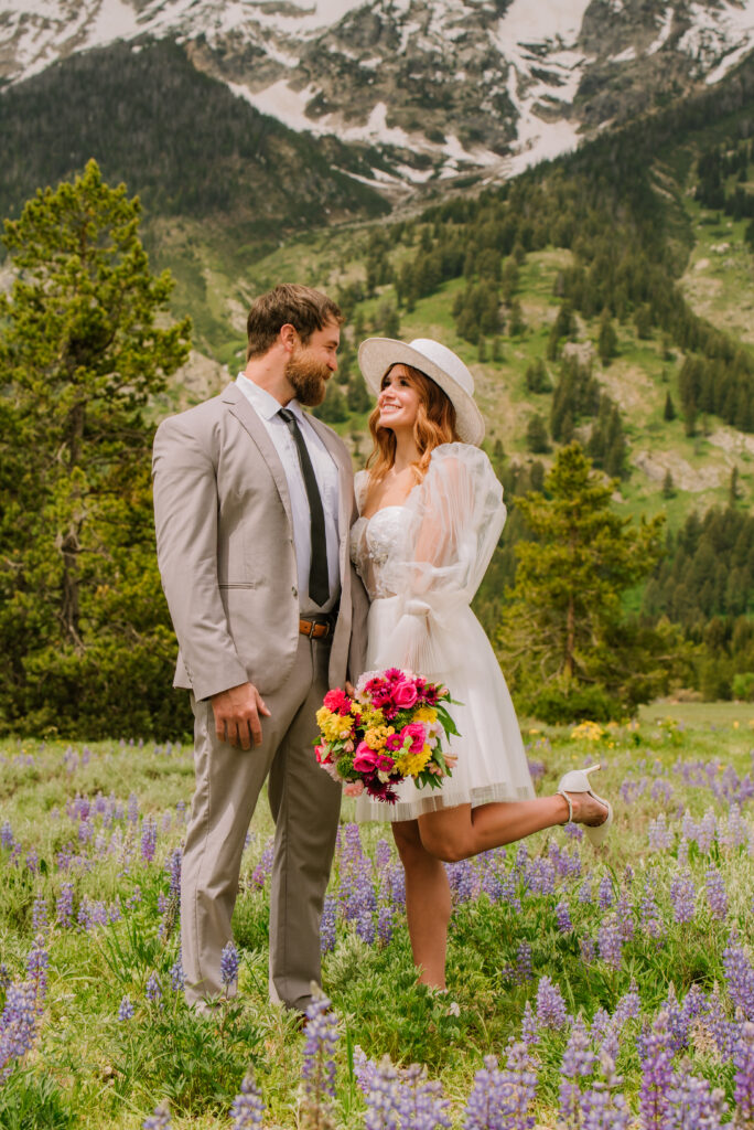 bride looks at groom while wearing a white cowboyhat, short dress, and bright colored bouquet in a field of wildflowers. 