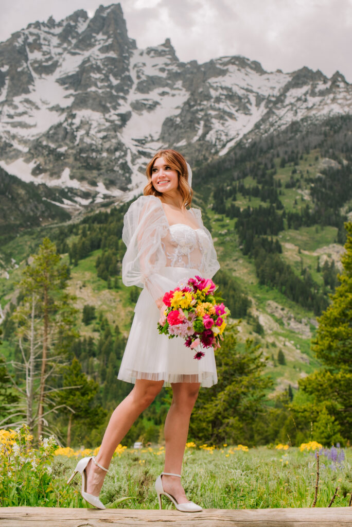 Bride in a field of wild flowers that are blue and yellow in grand teton nantional park wearing a short dress and veil.