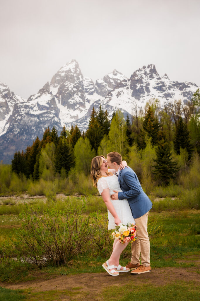 groom dip and kisses bride during Jackson Hole elopement. Bride is wearing short white wedding dress and white sandals. Groom is in tan pants and blue blazer