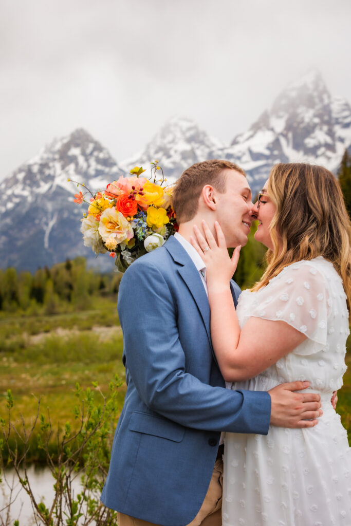 Bride holds her vibrant flowers at her minimalistic elopement in jackson hole while teasing groom to kiss her. Her hands are on his face and he is smiling while leaning in for a kiss.